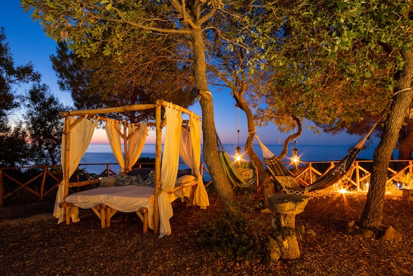 Una casa en árbol con cama y hamaca, rodeada de árboles y con vistas a un cuerpo de agua.