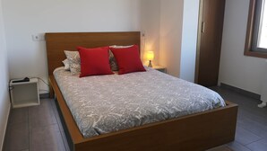 Uma cama de solteiro com colcha estampada e duas almofadas vermelhas, uma luminária de cabeceira e uma mesa de cabeceira com controle remoto.