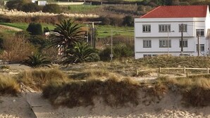 Uma praia arenosa com duas pessoas caminhando, um prédio branco com telhado vermelho ao fundo e uma palmeira.