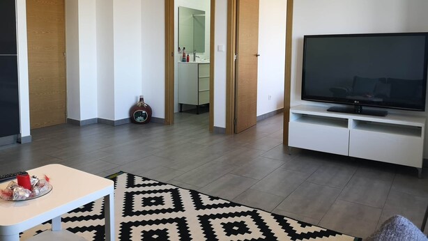 Uma sala de estar moderna com televisão de tela plana, um armário branco, um tapete estampado e uma pequena mesa com um objeto vermelho.