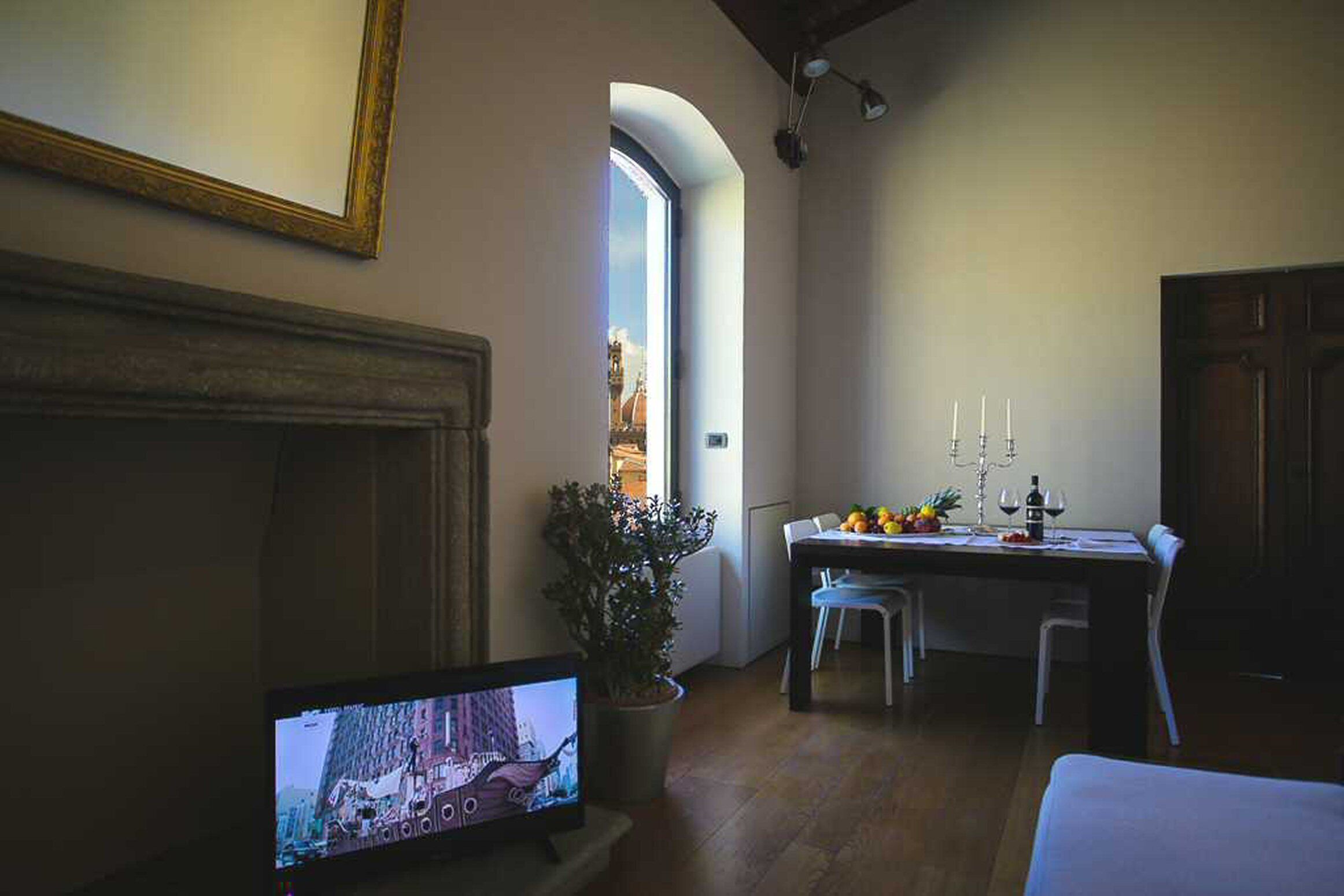 Comedor con una mesa para dos, una planta en maceta y un televisor mostrando un paisaje urbano.
