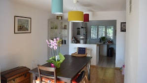 Un coin repas avec une table en bois, des chaises, un vase de fleurs et une vieille malle. On aperçoit une cuisine à l’arrière-plan, avec des placards et une fenêtre.