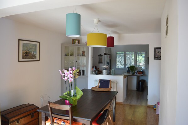 A dining area with a wooden table, chairs, a vase of flowers, and a vintage trunk. A kitchen is visible in the background with cabinets and a window.