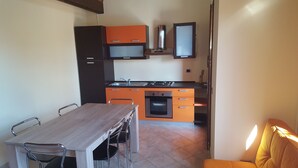 A compact kitchen with a dining area, featuring a wooden table and chairs, a built-in oven, and orange cabinetry. - Chalet in Sardinia (Villaputzu)