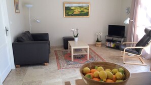 A living room with a sofa, a coffee table, a TV, and a chair. There is a painting on the wall and a bowl of fruit on the table.