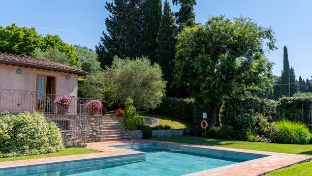A swimming pool with a stone deck, surrounded by lush greenery and a small building with a tiled roof.