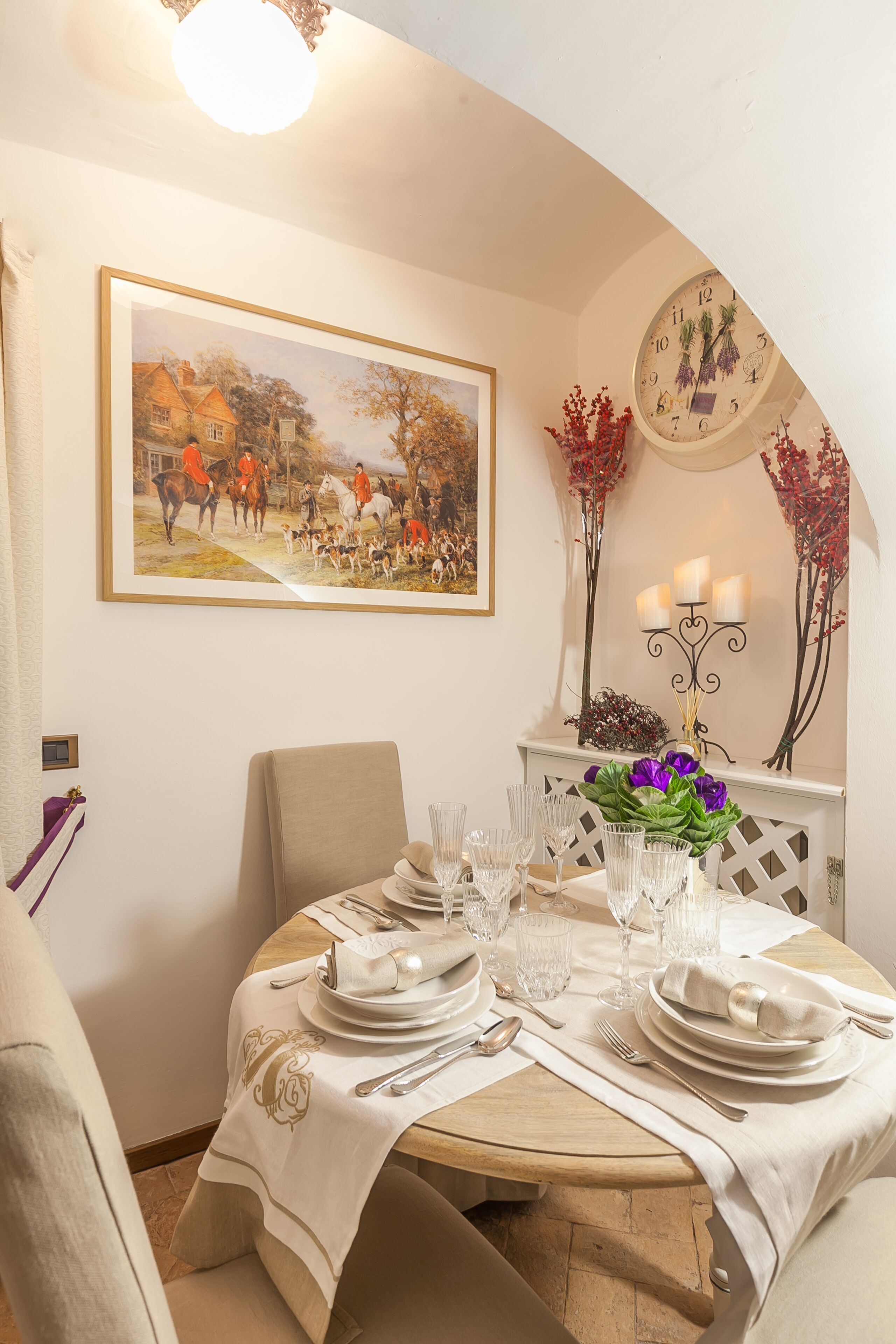 Un comedor con una mesa redonda dispuesta para una comida, un cuadro enmarcado arriba y un reloj fijado a la pared.