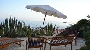 Un área de descanso frente a la playa con sillas de madera y una sombrilla blanca, con vista a una playa concurrida.