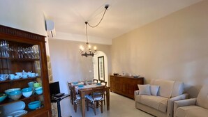 Comedor con mesa de madera dispuesta para cuatro personas, rodeada de sillas. A la izquierda, un mueble de madera con vajilla. En primer plano, un sofá beige, y al fondo, un mueble de madera con objetos decorativos.