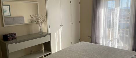 Une chambre à coucher comprenant un lit, une commode, un miroir, une armoire et une fenêtre avec des rideaux.