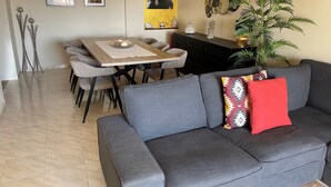 Un saló modern amb un sofà gris, taula de dinar, cadieres i una planta gran.