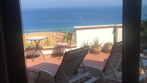 Un patio con dos sillones de descanso y una mesa, con vistas a la costa, el mar y un barco a lo lejos.