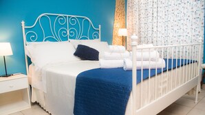 A neatly made bed with white linens and a blue quilt, a bedside table with a lamp, and a patterned curtain. - Smell of sea house (Avola)