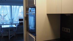 Cozinha moderna com armários brancos, bancada preta e pia de aço inoxidável. É possível ver uma área de refeições ao fundo, com uma mesa e cadeiras.