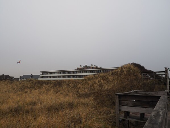 A modern building with a flat roof and multiple windows, surrounded by tall grass and a wooden structure.