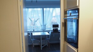 Corredor estreito que dá para uma sala de jantar com mesa e cadeiras, uma janela com cortinas finas e um forno embutido.