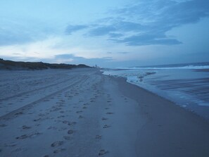 A beach with footprints leading to the water, a dune, and a distant structure.