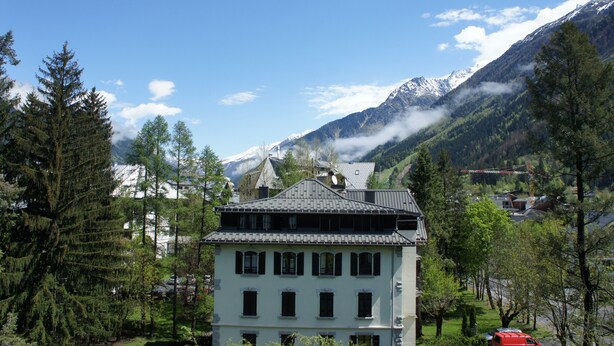 Un bâtiment à deux étages, doté d’un toit en pente, entouré d’arbres et avec des montagnes en arrière-plan.