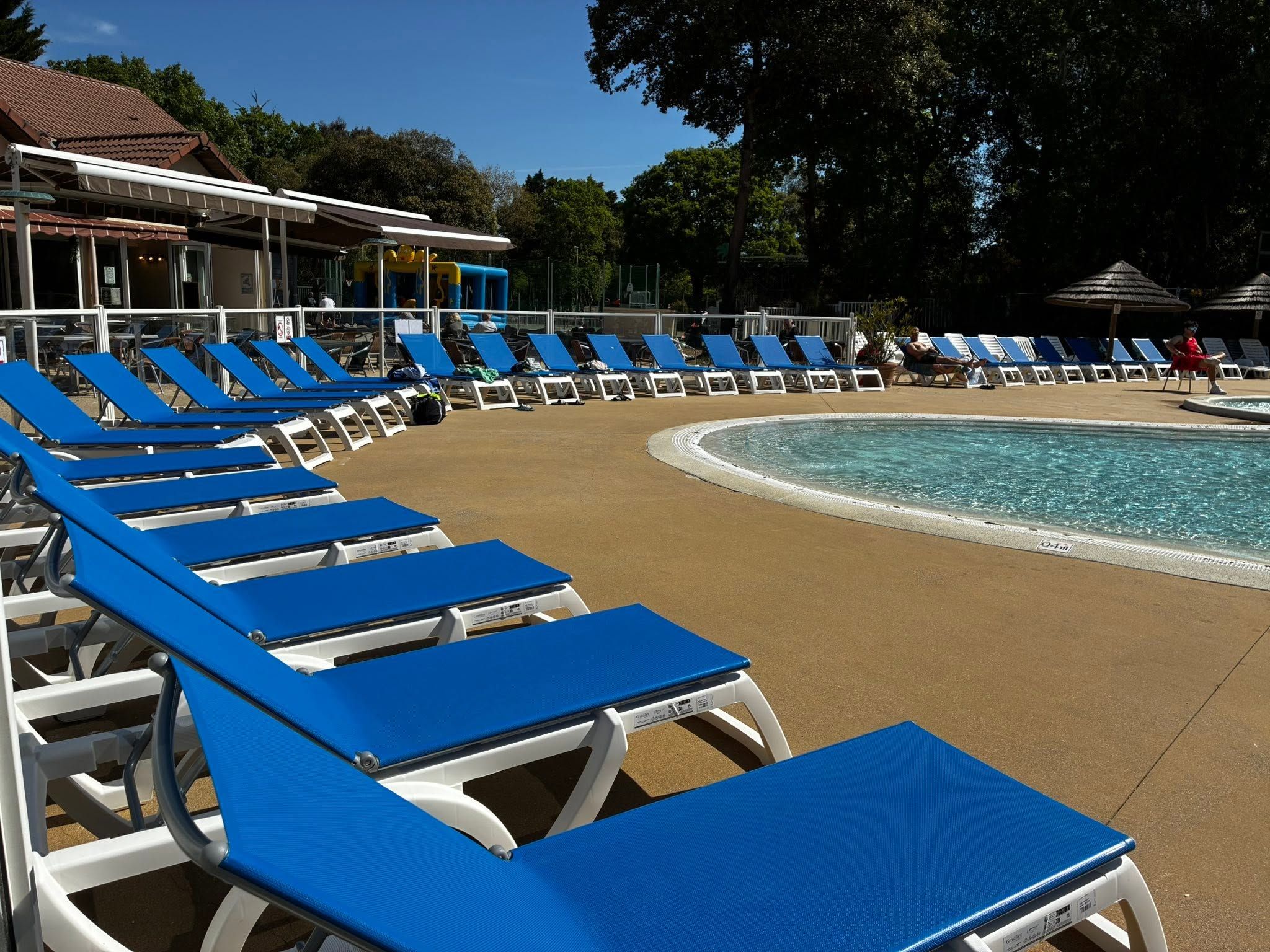 Un espace aménagé au bord de la piscine, avec des chaises longues bleues disposées en rangées, une piscine à l’eau claire et, au fond, un bâtiment doté d’un toit recouvert de tuiles.