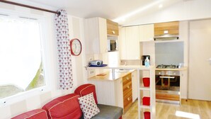 Un espace de vie compact comprenant un coin cuisine, un canapé avec des coussins rouges, un rideau à motifs et une horloge au mur.