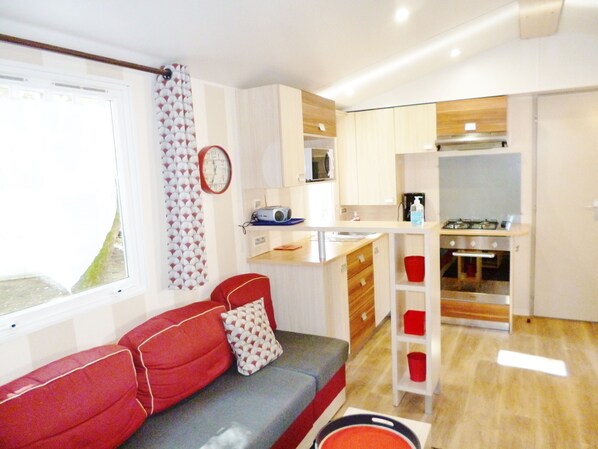 A compact living space with a kitchenette, a sofa with red cushions, a patterned curtain, and a clock on the wall.