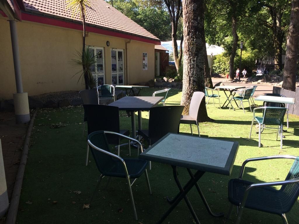 Ein Außenbereich mit Terrassentischen und -stühlen, ein Gebäude mit rotem Dach und Bäume.