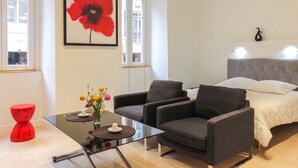 A modern room with a bed, two armchairs, a glass coffee table, and a red stool. There is a large red flower painting on the wall and a window with a view of buildings. - Studio charm in the heart of Old Nice (Nice)