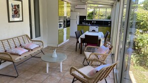 A sunlit patio with wicker furniture, a small table, and a kitchen area visible through glass doors. - House with small garden in a quiet area 3 minutes from the town center (La Rochelle)