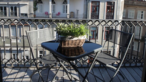 Un balcón con una mesa, sillas, una planta en maceta y vista a un edificio histórico con cúpulas y una cruz.