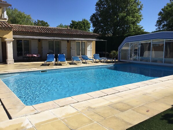 A swimming pool with lounge chairs, a covered area, and a building with a tiled roof.