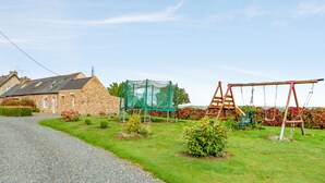 A rural setting with a stone house, a gravel path, a swing set, and a green netted area.