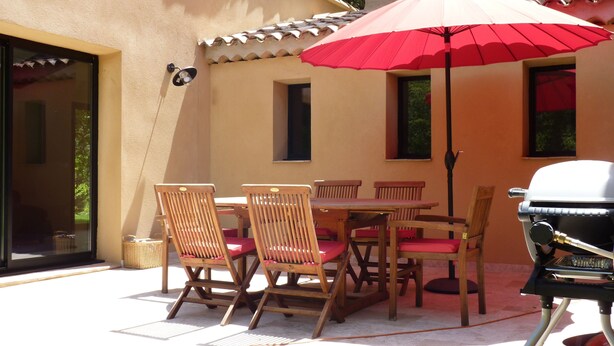 Une terrasse extérieure avec des chaises et une table en bois, un parasol rouge et un barbecue.