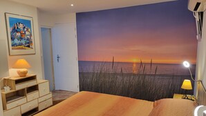 Une chambre à coucher comprenant un lit, une table de chevet, une lampe et une fresque murale représentant un coucher de soleil sur la mer.
