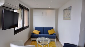 Un espace de vie compact comprenant un canapé, deux fauteuils, une table basse en verre, un téléviseur à écran plat et un tableau accroché au mur.