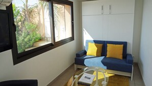 Un salon avec un canapé bleu, des coussins jaunes, une table basse en verre avec une télécommande, et une fenêtre donnant sur un espace verdoyant.