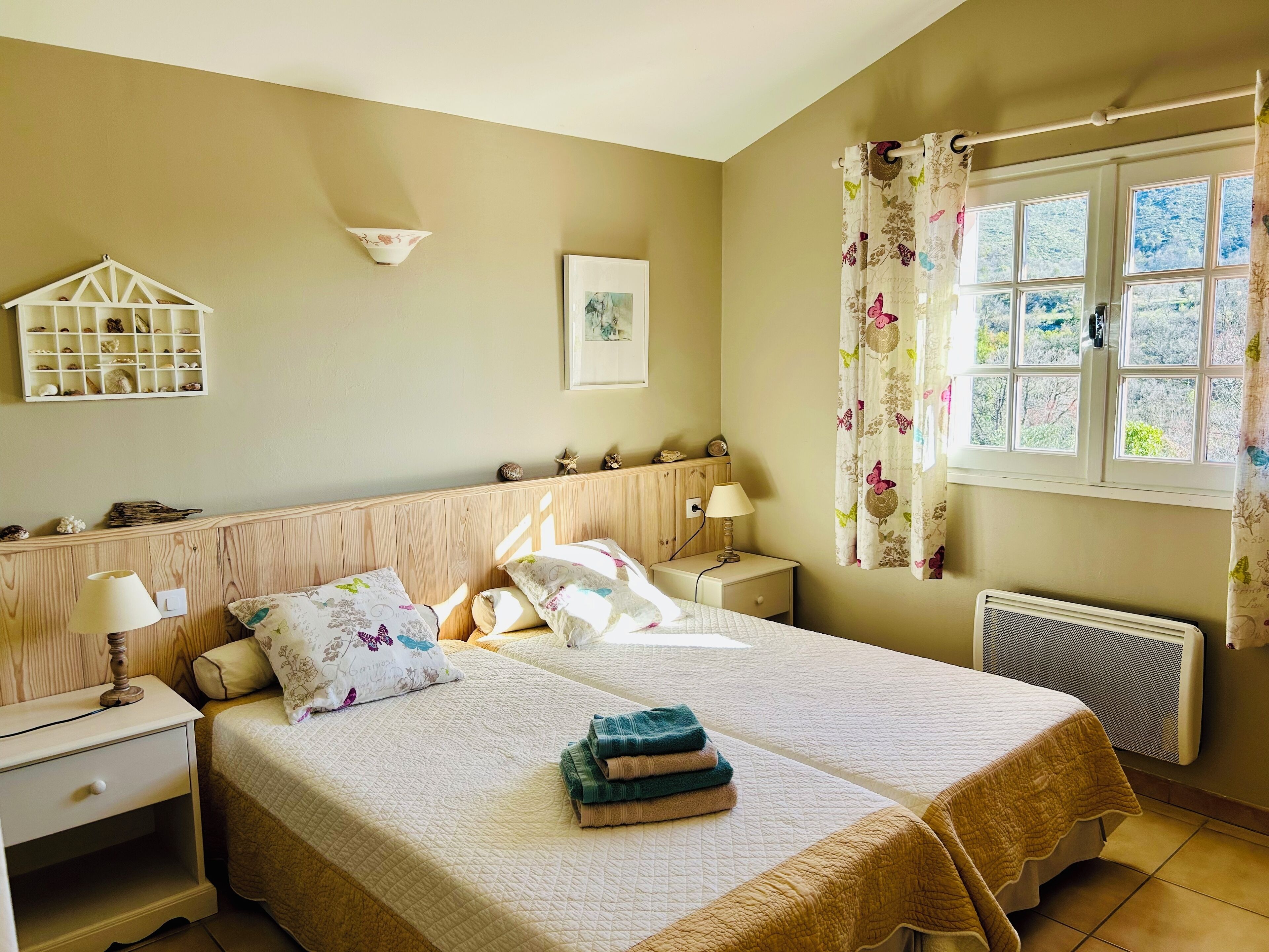 Un dormitorio con cama, dos mesitas de noche, lámpara, estante en la pared con objetos decorativos, ventana con cortinas y radiador.