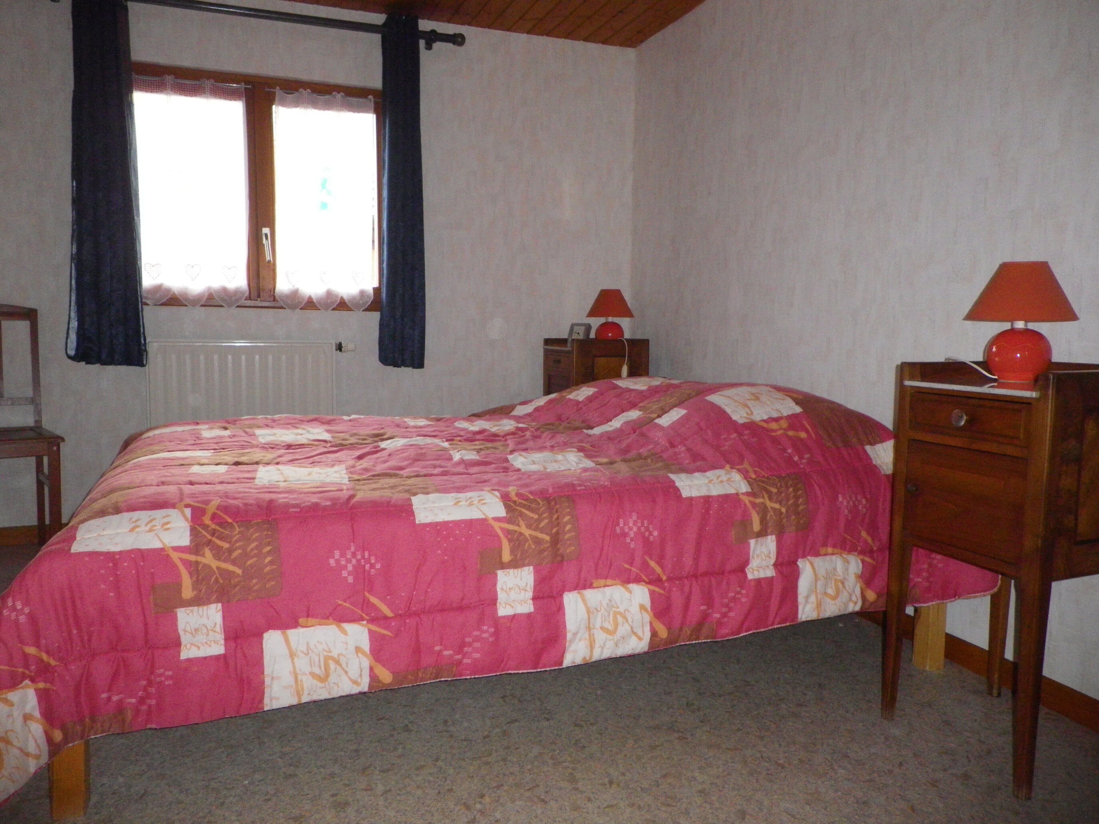 Ein Schlafzimmer mit Bett, Nachttisch, Kommode, einem Fenster mit Vorhängen und einer Heizung.