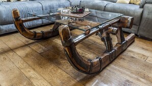 Una mesa de centro de madera con superficie de vidrio, ubicada frente a un sofá gris con cojines estampados.