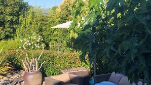 An outdoor seating area with wicker furniture and a glass-top table, surrounded by greenery and a large potted plant. - Air-conditioned villa with private pool and jacuzzi, in a quiet location close to beaches. (Appietto)