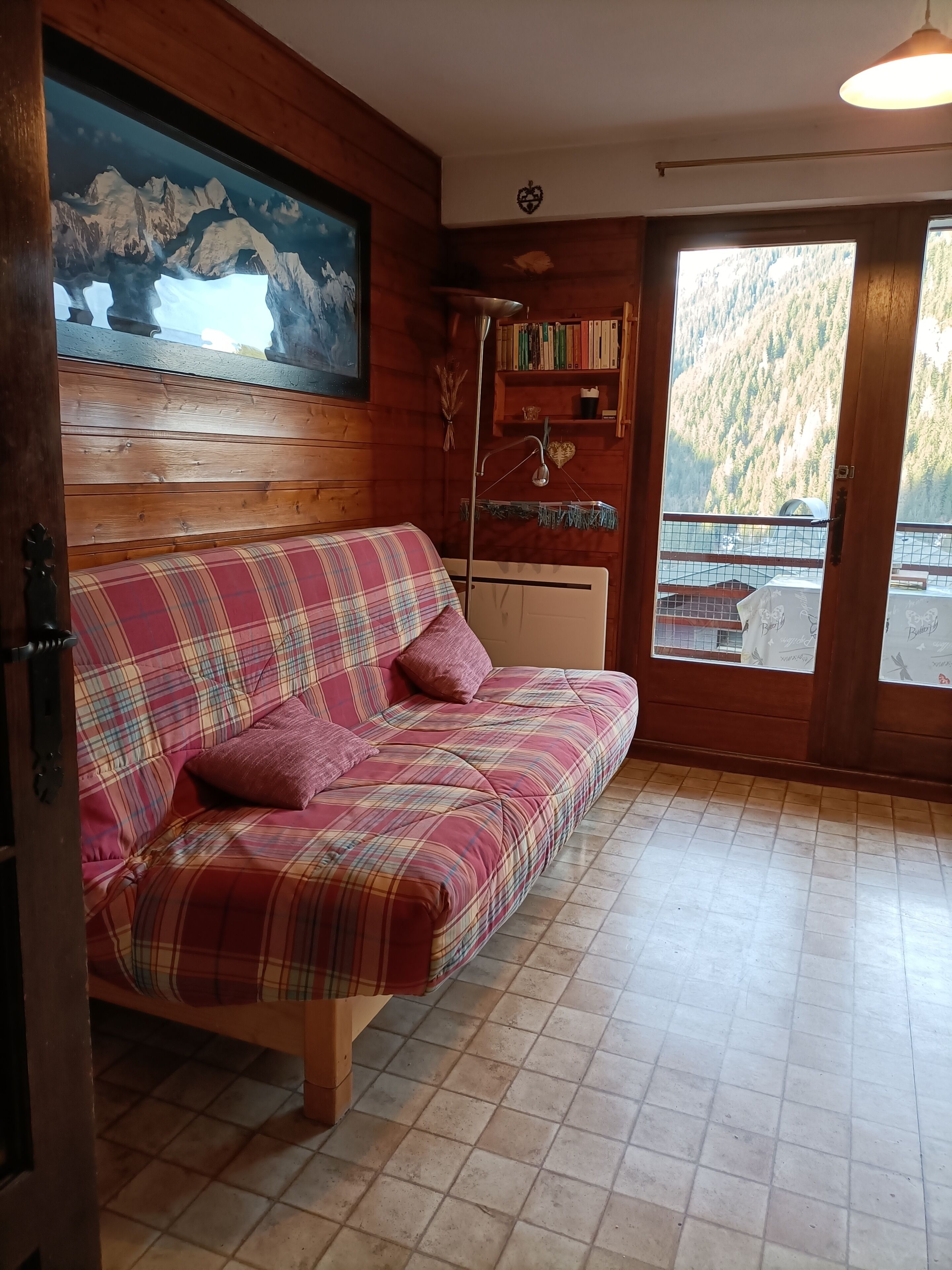Une pièce chaleureuse avec un canapé à carreaux, une bibliothèque, un tableau représentant des montagnes et une vue sur une forêt, visible à travers une grande fenêtre.