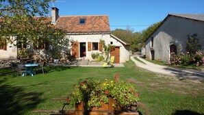 A rustic stone house with a tiled roof, a garden with potted plants, and a gravel path.