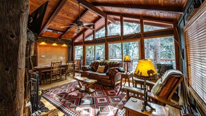 A cozy cabin interior with a wooden ceiling, large windows, a fireplace, and a patterned rug.