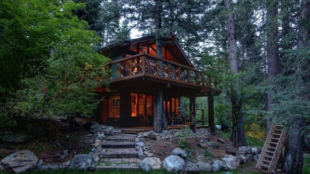 A rustic cabin with a wooden deck, surrounded by a stone pathway and a wooden ladder.