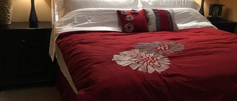 Un lit bien fait, recouvert d’une couverture rouge et agrémenté d’oreillers blancs, avec deux lampes de chevet de chaque côté et des œuvres d’art encadrées accrochées aux murs.