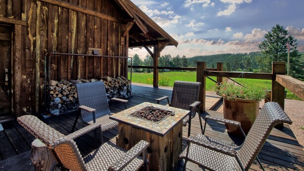 An outdoor deck with wicker furniture, a fire pit, and a view of a green field. - Beautiful Timber Frame Canyon with Spring Creek Canyon views! (Rapid City)