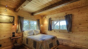 A wooden cabin bedroom with a bed, windows, curtains, a nightstand, and a framed map on the wall. - Beautiful Timber Frame Canyon with Spring Creek Canyon views! (Rapid City)