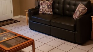 A living room with a sofa, patterned pillows, a glass-top coffee table, a lamp, and a painting of a waterfront scene.