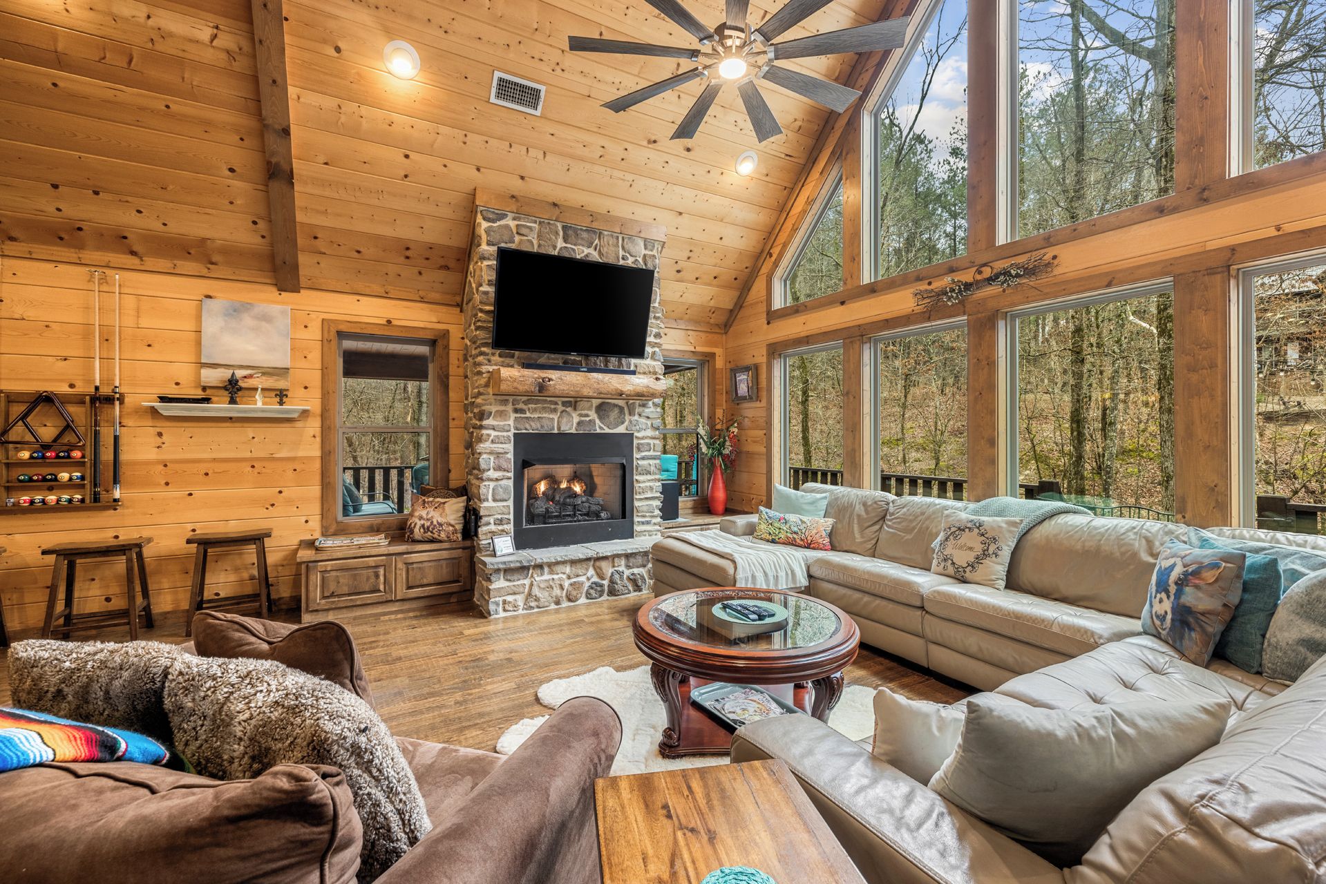 A cozy cabin interior with a stone fireplace, wooden walls, and large windows offering a view of the forest.