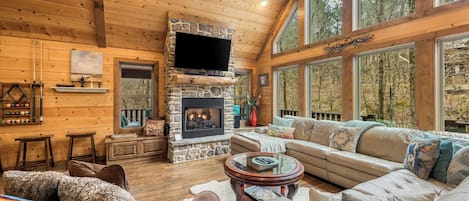 A cozy cabin interior with a stone fireplace, wooden walls, and large windows offering a view of the forest.