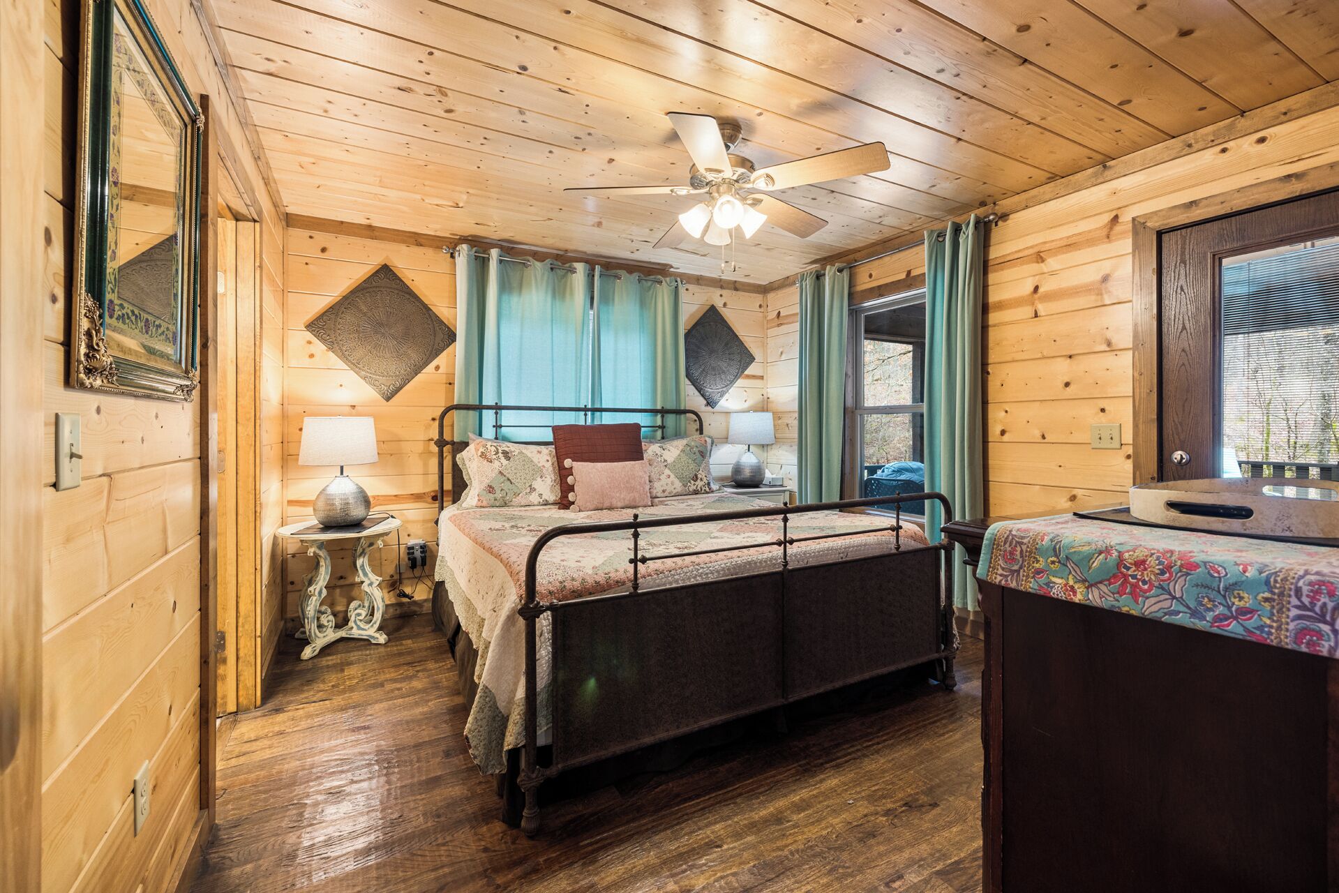 Un dormitorio de cabaña de madera con cama, dos mesitas de noche, un ventilador de techo y una ventana con cortinas.